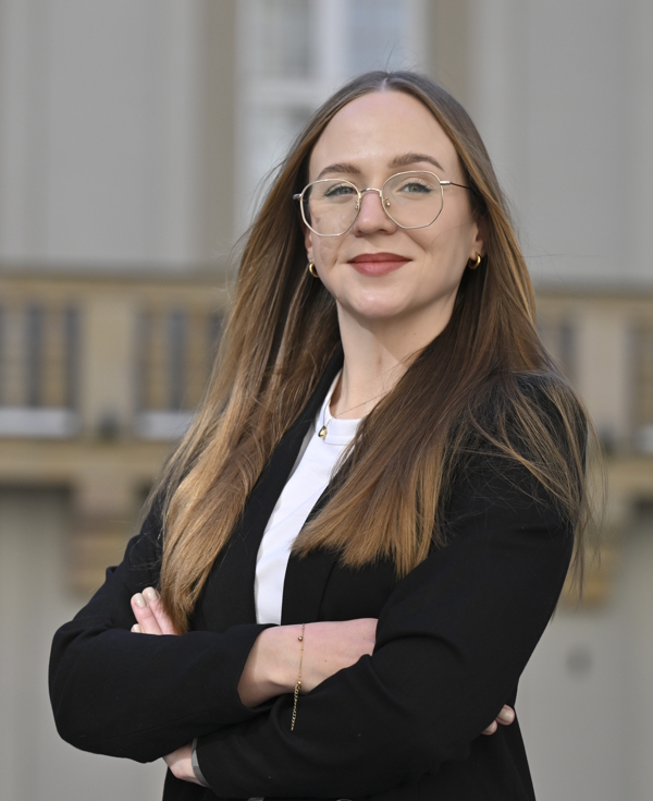 Portret uśmiechniętej kobiety z długimi jasnymi włosami w okularach i ciemnej marynarce na tle budynku.