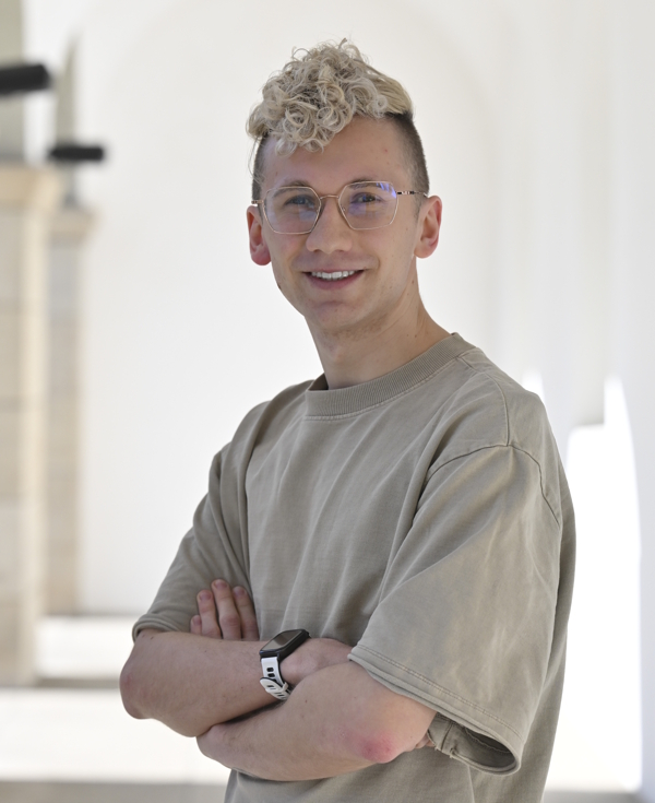Młody mężczyzna z blond kręconymi włosami i okularami uśmiecha się, stojąc z założonymi rękami.