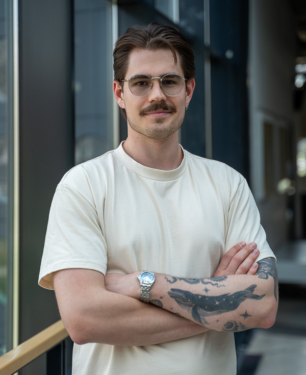 Mężczyzna z tatuażami i w okularach, stojący na tle okien budynku.